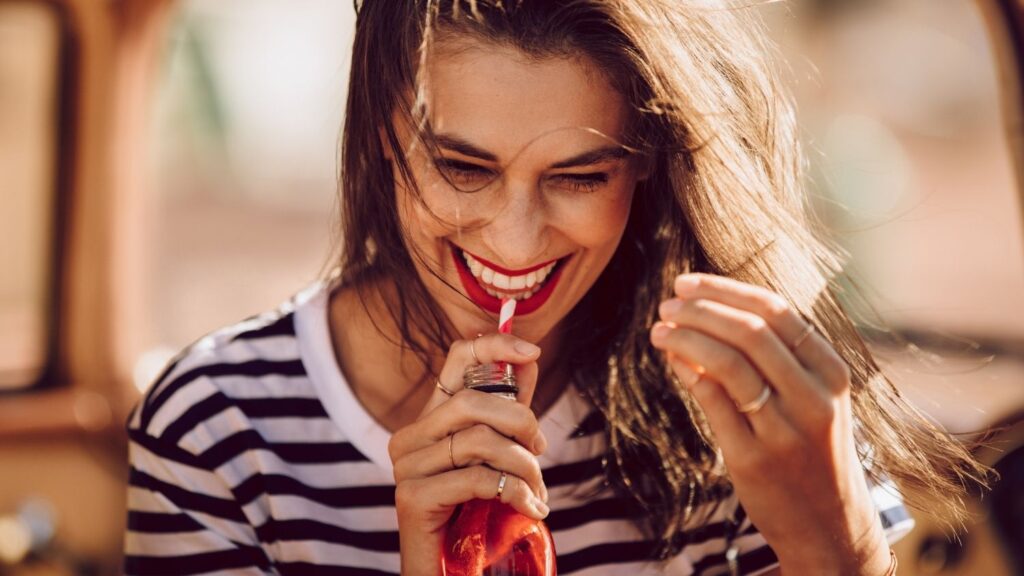 Woman smiling and drinking from a glass bottle outdoors.